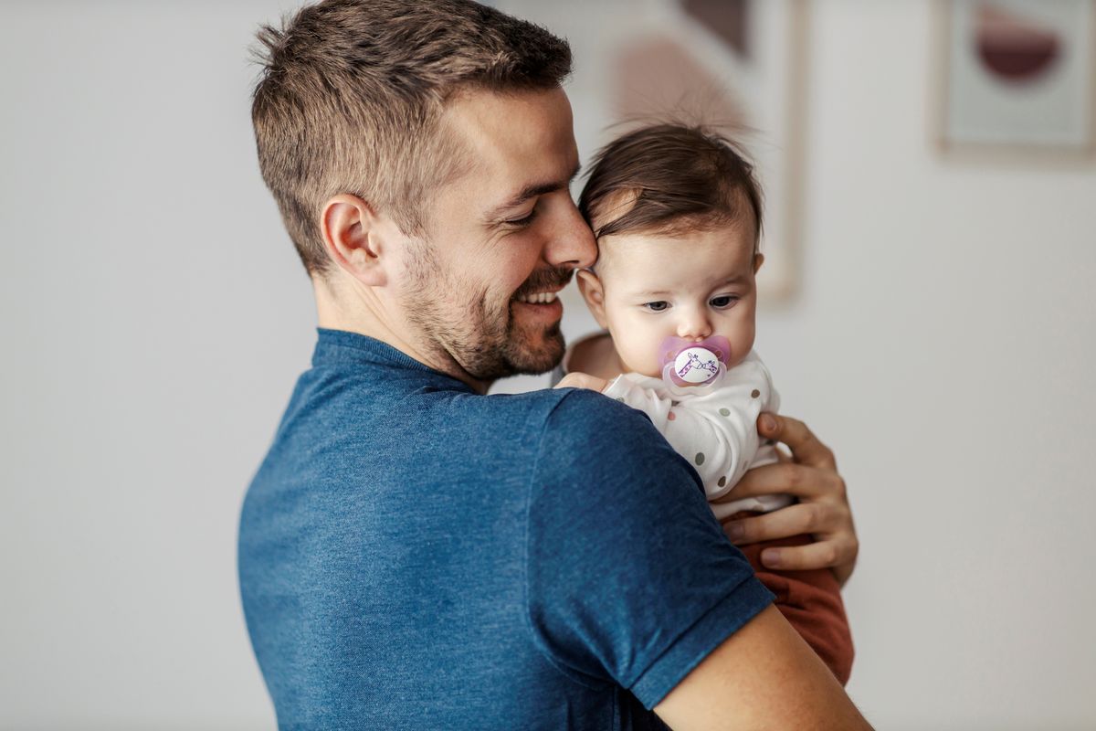 Pai segurando a filha bebê no colo, encostando o rosto no dela, em ambiente doméstico de fundo claro