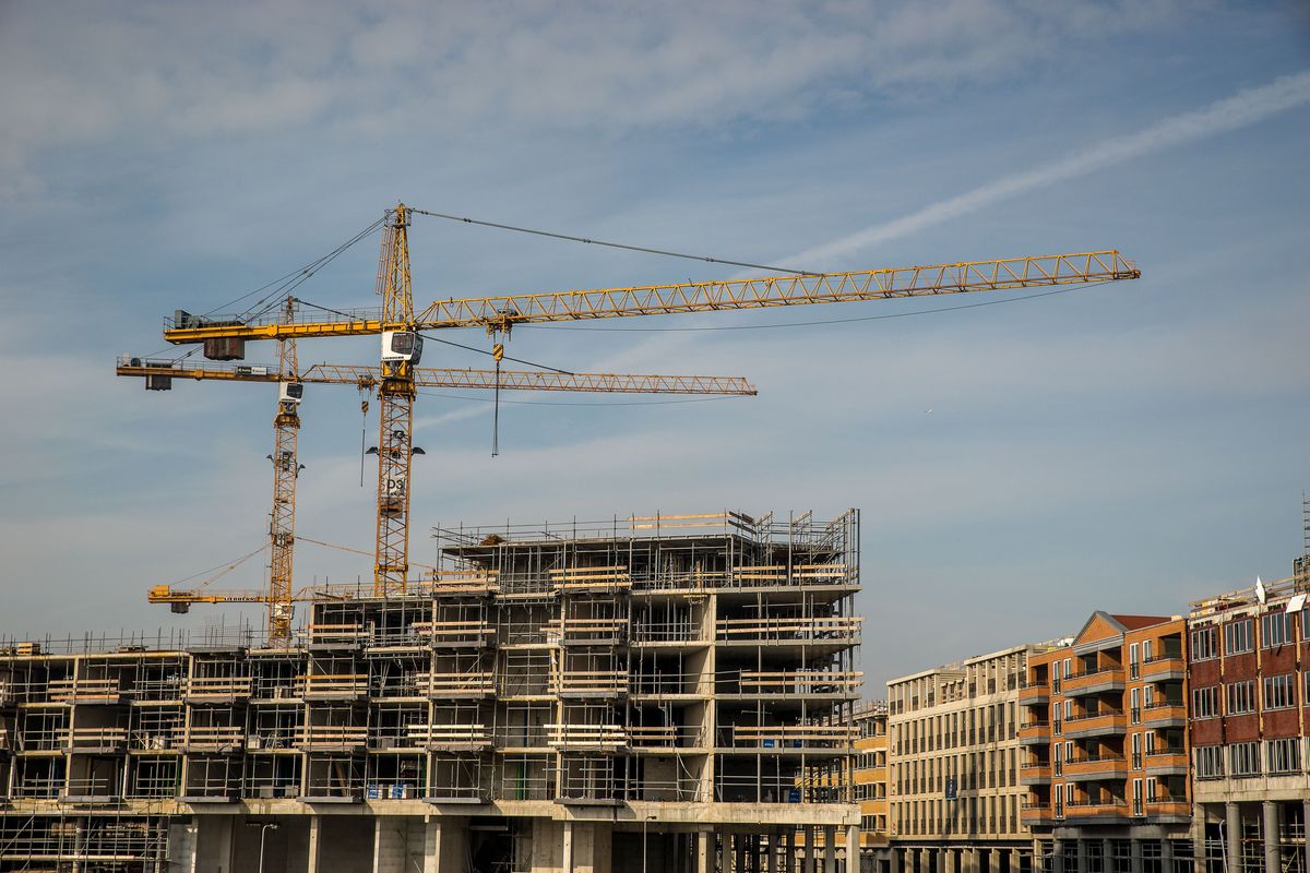 Canteiro de obras com três guindastes amarelos sobre a estrutura de concreto de um prédio residencial em construção, com edifícios de tijolo ao lado e céu azul